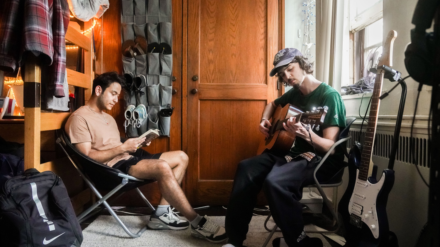 Two students relax in a dorm room, one reading and one playing acoustic guitar