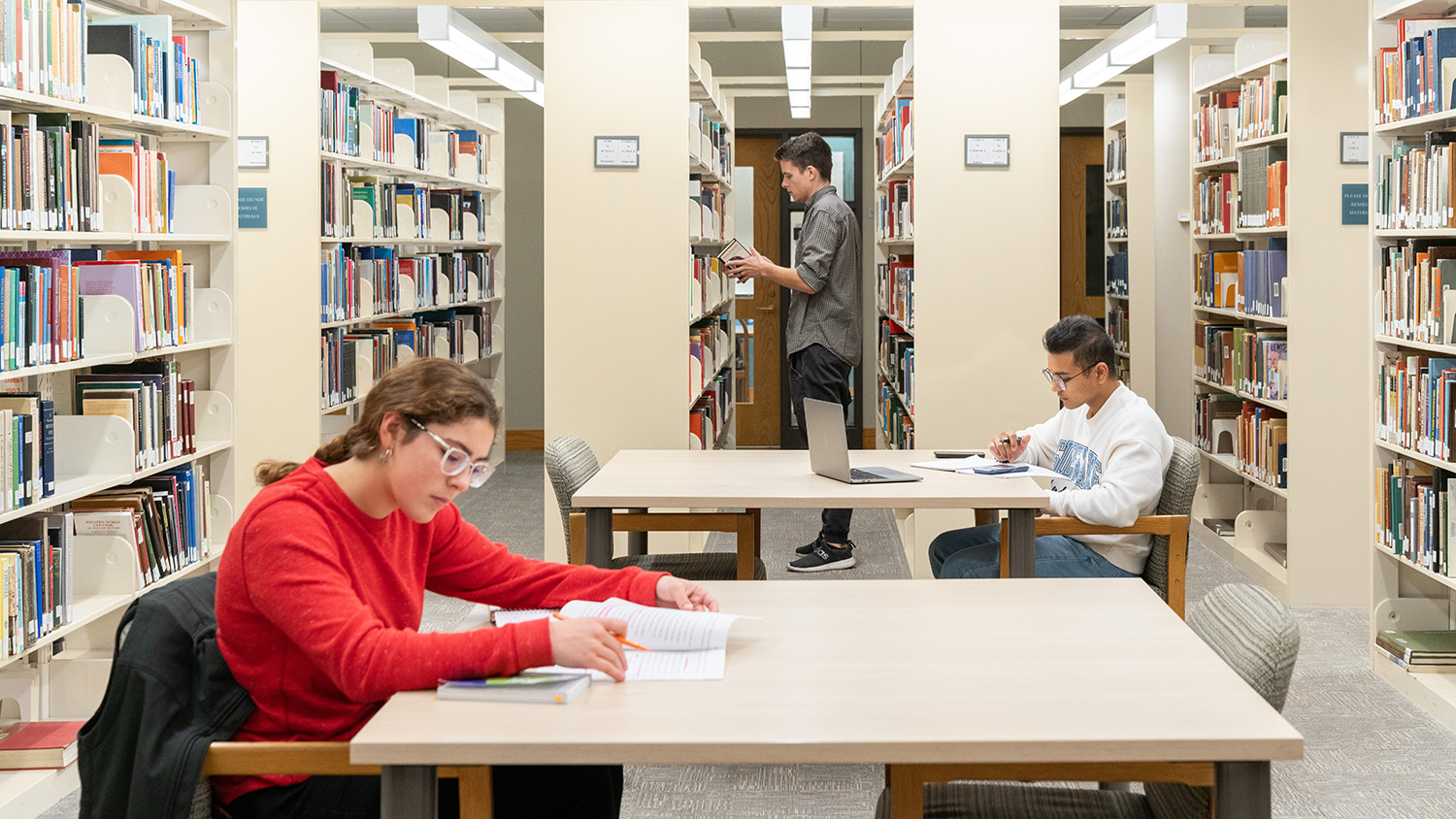 Three students are in the library. One is looking at books, and the other two are studying independently at tables.