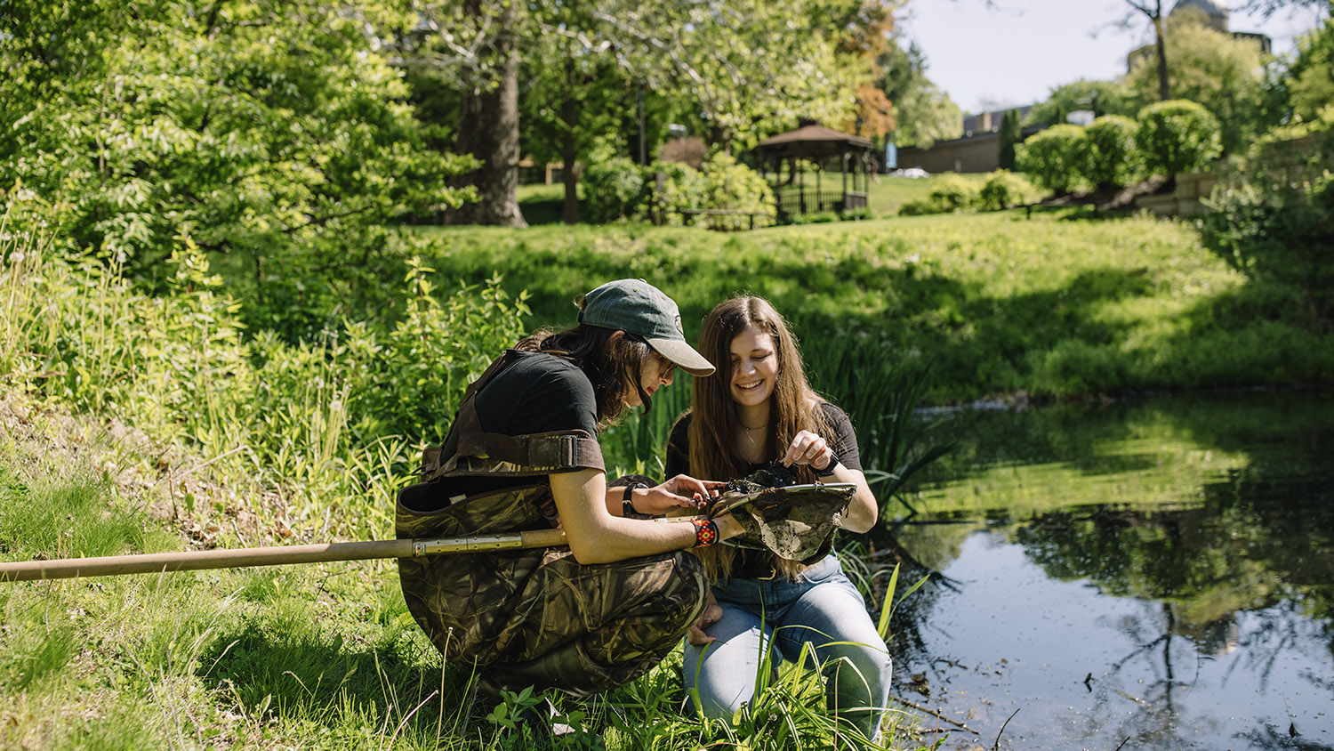 Two students explore pond life near campus. One is wearing waders.