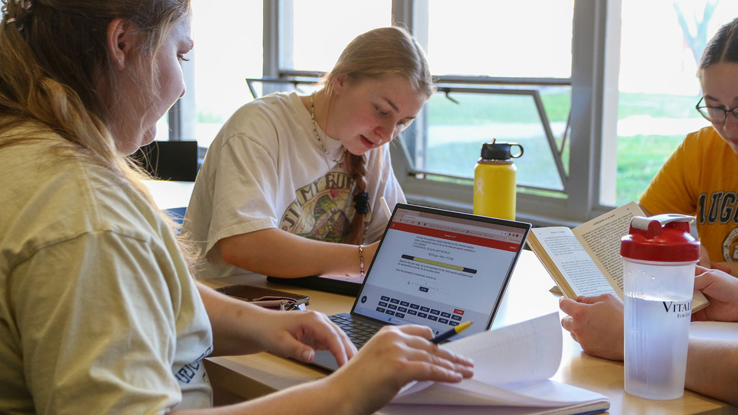 Three students study at a table near a window in a residence hall lobby