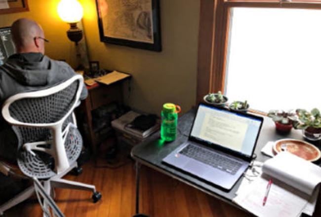 Man in the corner on computer and laptop on a desk. 3 small potted plants on the table in front of the window.