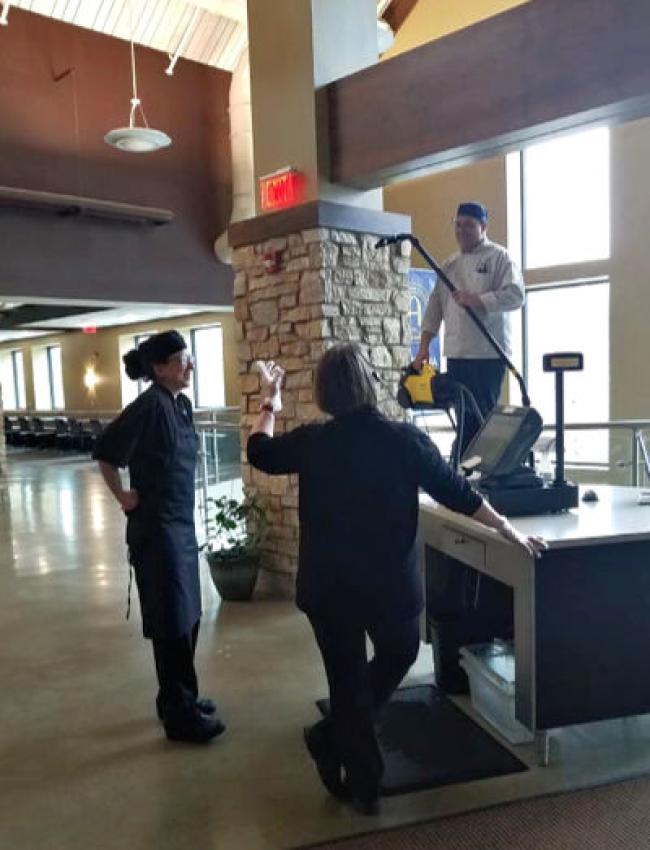 Kevin standing on a table holding a hand held vaccum while talking to Katie and Kris