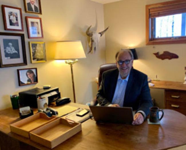 President Bahls sitting in his office chair behind his desk. Photos on the wall and a lamp in the corner.