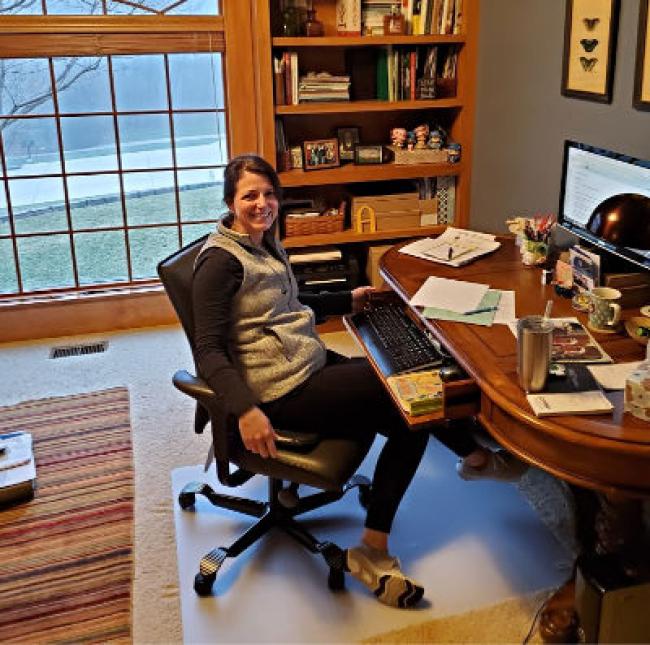 Sheri Curran sits at desk with computer and keyboard in lap. Books in the background and big bay window with lot light in the back.