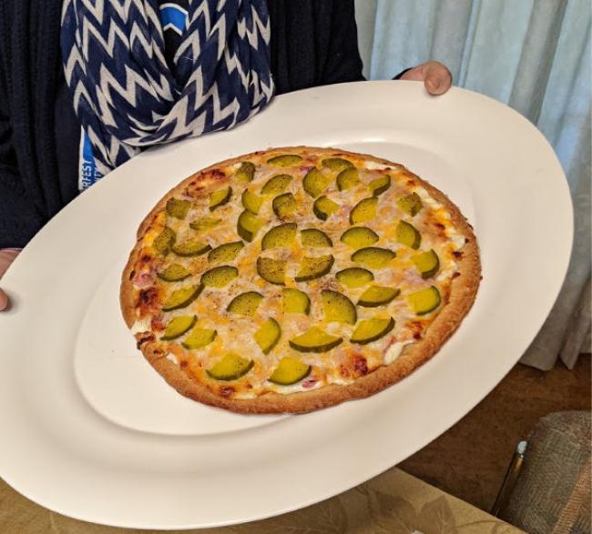 Heather holding a homemade pizza with green pickles on top a bed a melted cheese.