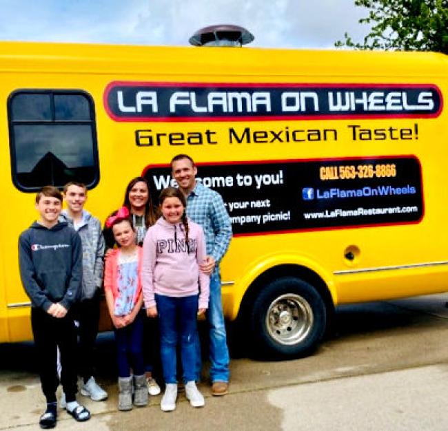 Ashley standing in front of a yellow taco truck to the bottom left of the photo. With her is her 2 sons, 2 daughter and husband.